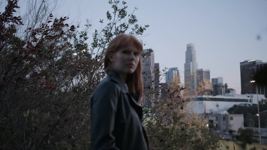 A woman stands near bushes with a city skyline behind her