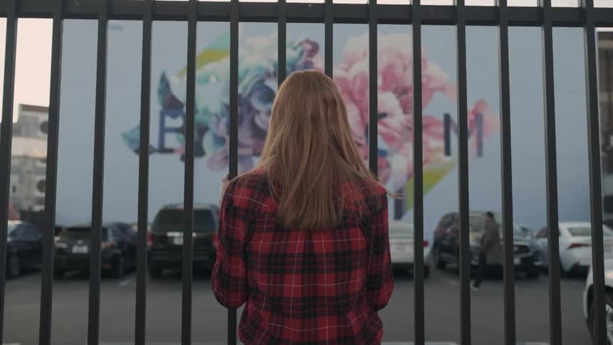 A woman is standing at a fence looking across a parking lot towards a mural