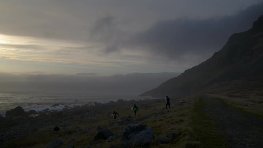 Men carrying surfboards hike along a rugged coastline
