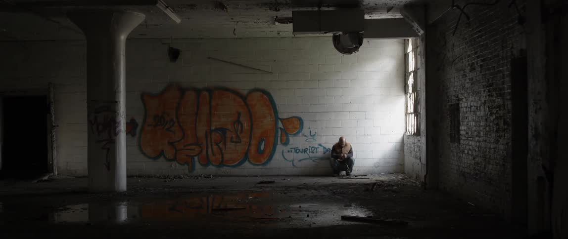 A man is sitting in a dark warehouse by the window