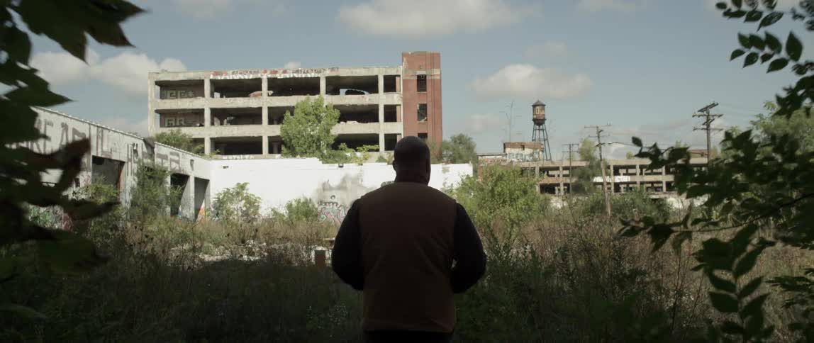 A man walks out through a tunnel into an overgrown lot during the day