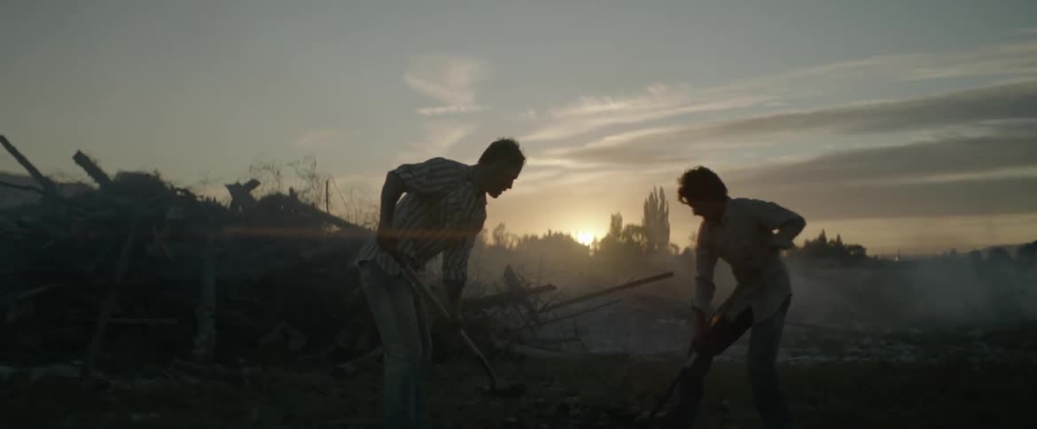 Two men are shoveling dirt on a pile of smoldering wood at dawn
