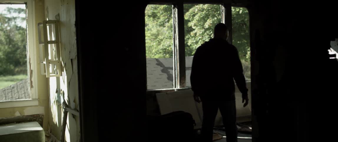 A man walking through a run down building stops to look out of a window