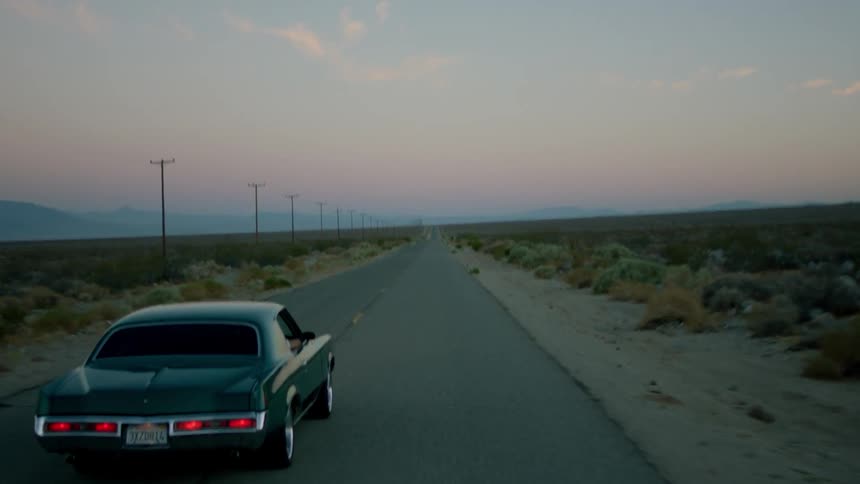 A late model car is being driven down a desert road at dusk