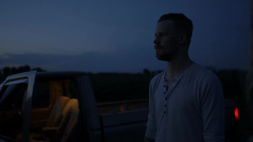 A man and woman are standing staring ahead outside a pickup truck on a rural road at dusk