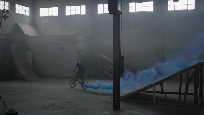 Man does a backflip on a bicycle with blue smoke trailing behind him