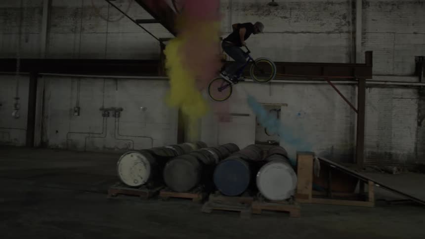 Man jumping and spinning on bike over barrels shooting up colored powder