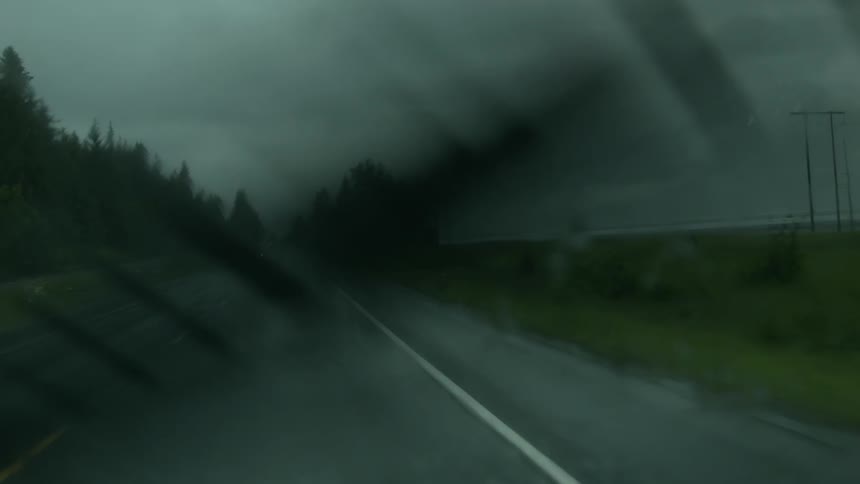 A vehicle's windshield wipers swipe back and forth as it drives on a road during a rainstorm