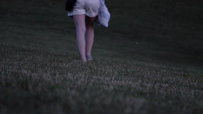 A woman is walking barefoot across a field with fireflies are flying around