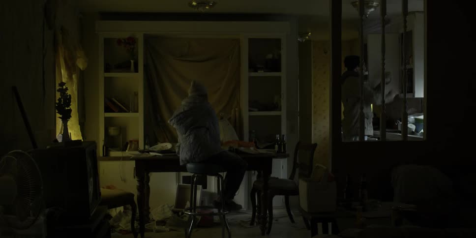 Man sits at desk while another man is in the kitchen