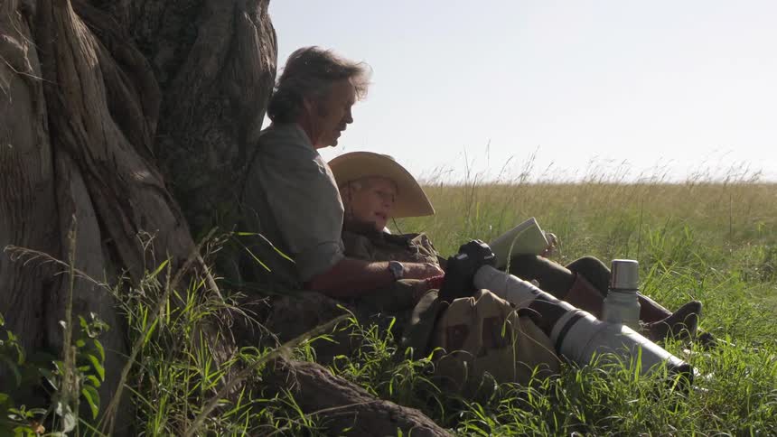 A couple sit against a tree while reading a  book