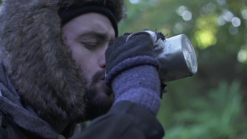 A bearded, brunette man wearing a fur hat and taking a drink from a thermos