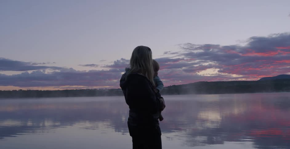 A woman and infant stand on a dock by a lake