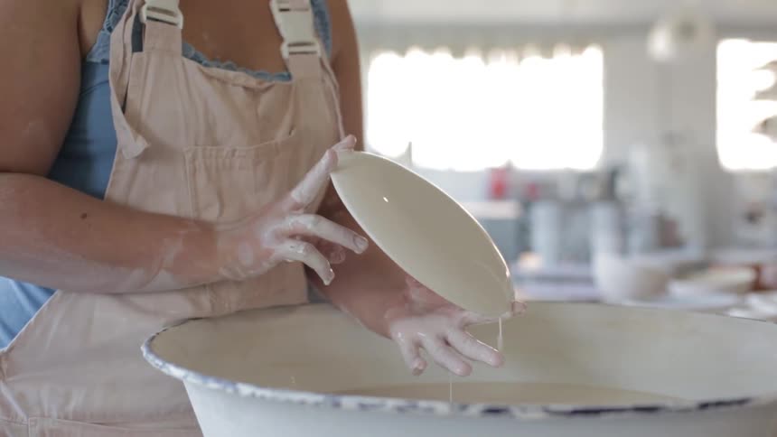 A woman in a sunlit ceramics workshop glazes a plate