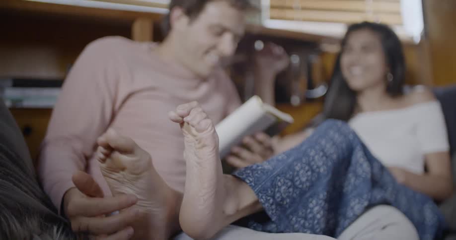 A man tickles a woman's feet while he reads in bed with her