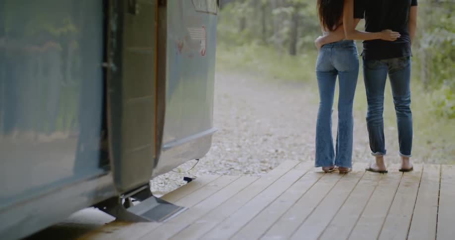 A man and woman stand with their arms around each other on a deck in front of a camper trailer