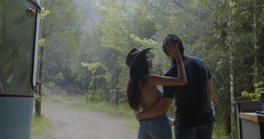 A woman caresses a man's head as they stand outside during the day