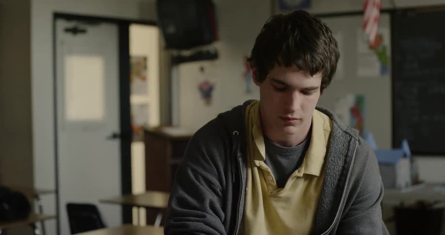 A young man is sitting in a classroom looking down at something