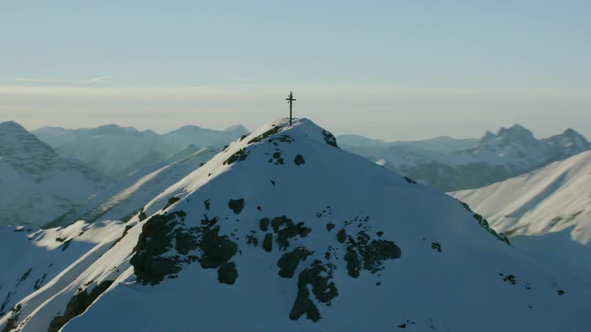 A cross stands atop a mountain peak covered in snow on a sunny day