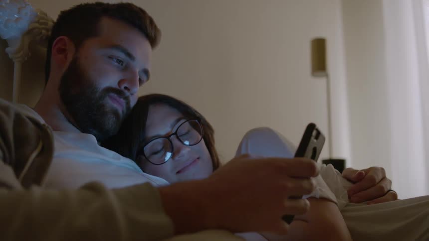 Man and woman cuddle in bed and look at a phone