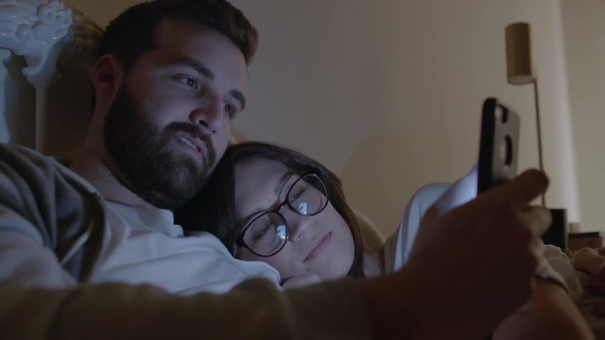 Man and woman snuggle in bed while looking at a phone
