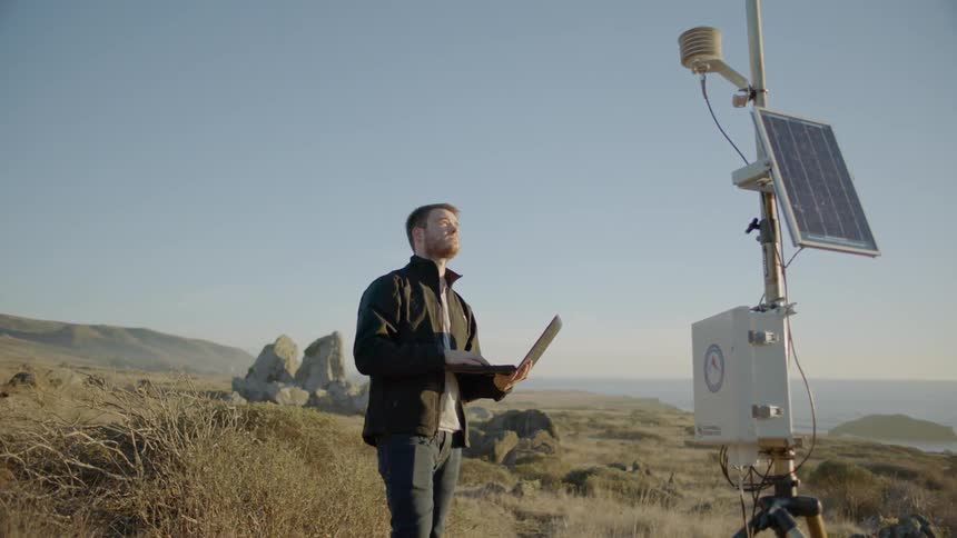 A man stands by a weather monitoring device with a laptop