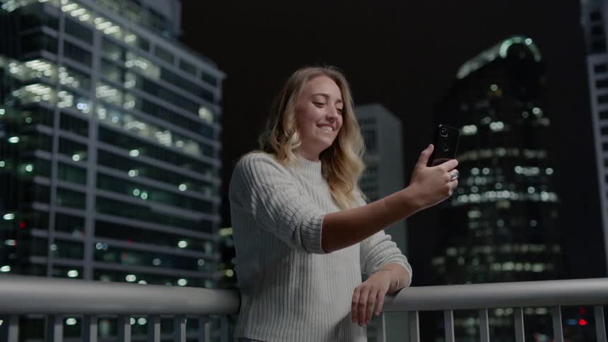 A woman is laughing and taking a photo of herself and the city skyline at night