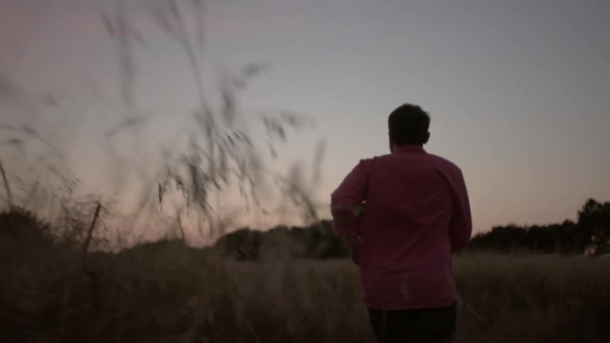A young white man walks through a grassy field next to a fenced in pasture in a rural area