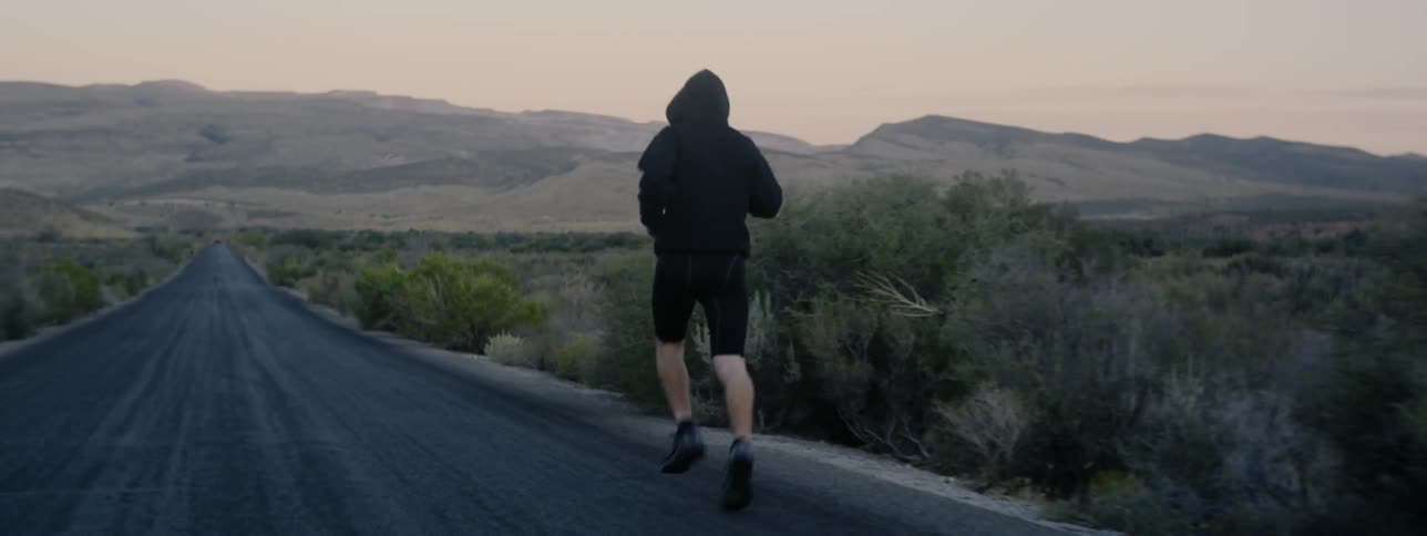 A man in a hoodie and shorts is jogging down a desert highway