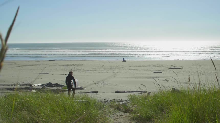A man walks slowly towards the water with a surfboard