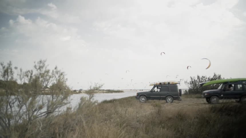 Two jeeps pull up to the water line and others are flying kites in the background
