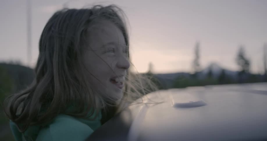 A young girl smiles while hanging out of a running car