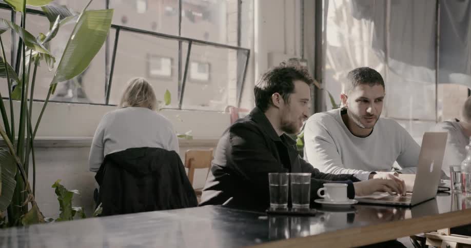 Men sitting in a cafe talk and look at a laptop as one types on the laptop