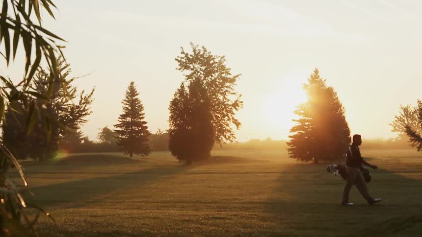 Man carries his golf clubs across a course at sunset