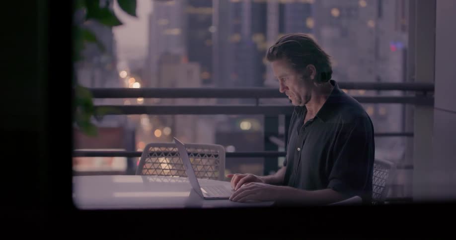 Man sitting at table on balcony with city view is typing on laptop