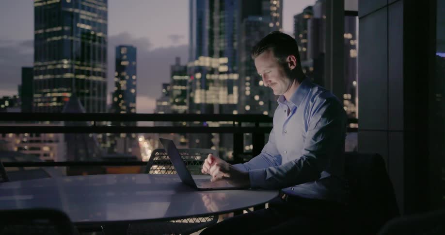 A man is working on his laptop with a huge window over his shoulder of a big city