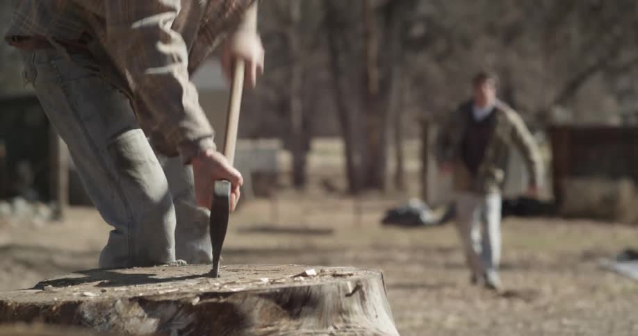 A man approaches another who is splitting logs outside