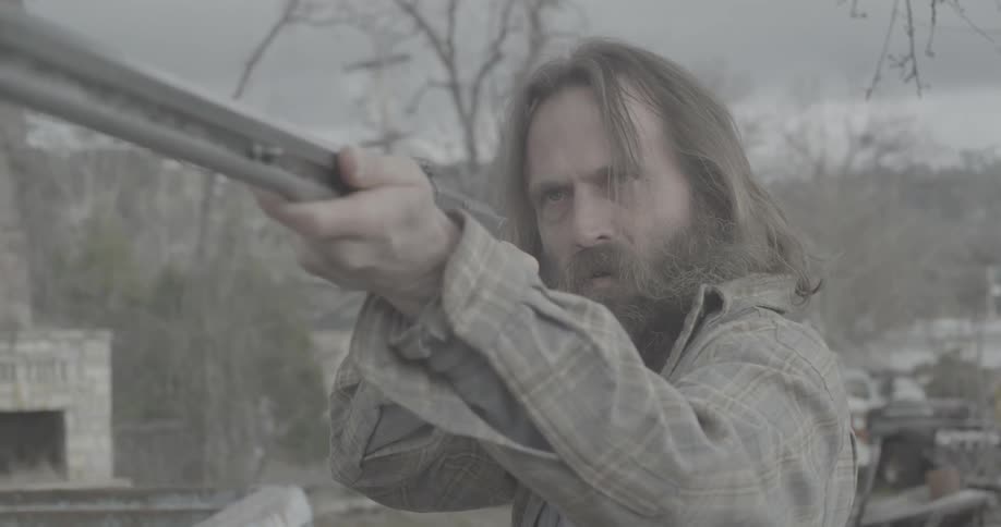 A bearded man points a shotgun angrily