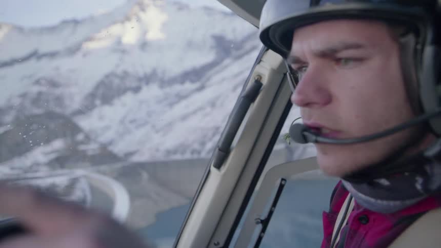 A pilot flies a helicopter over snowy mountains