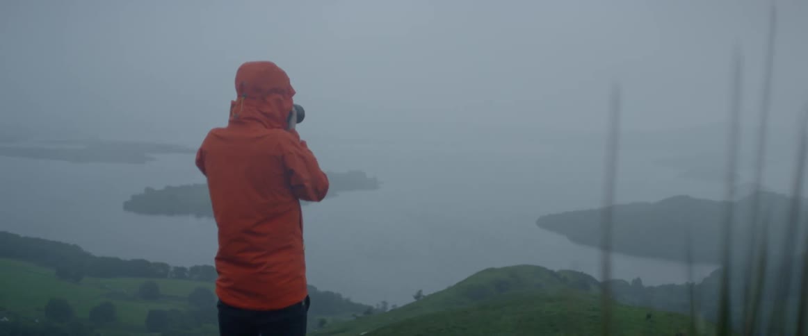 Man wearing an orange rain coat takes pictures of a body of water while standing on a hilltop