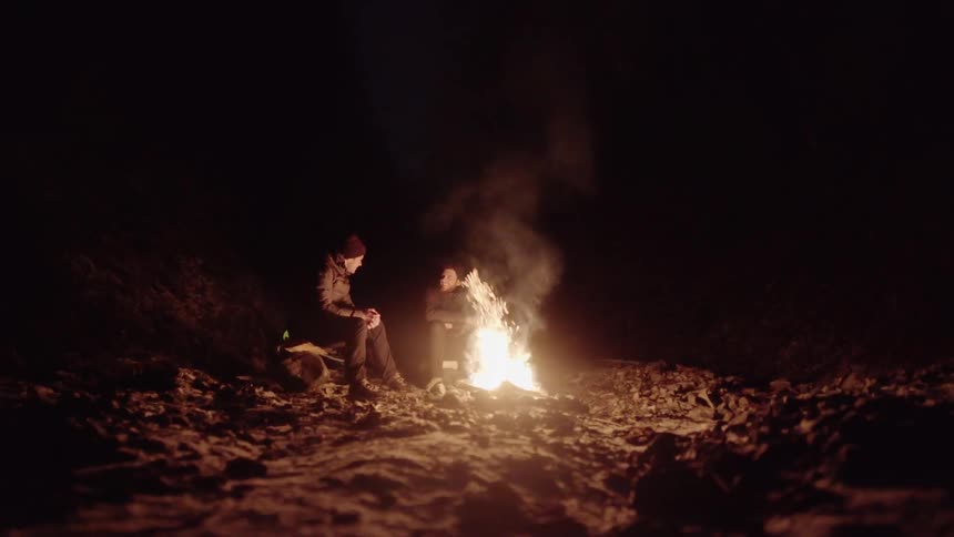 Two men are sitting by a fire at night