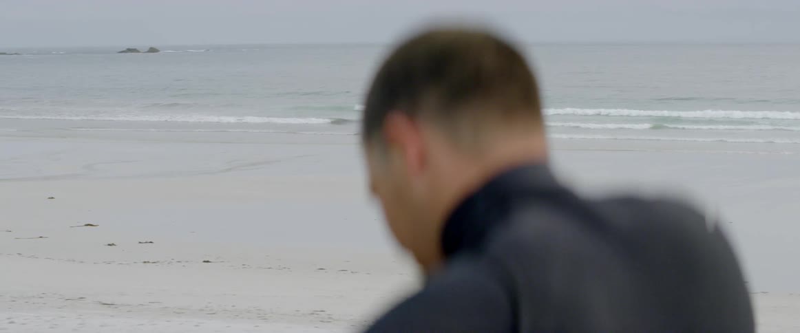 Man wearing a wet suit looks down while standing on a beach