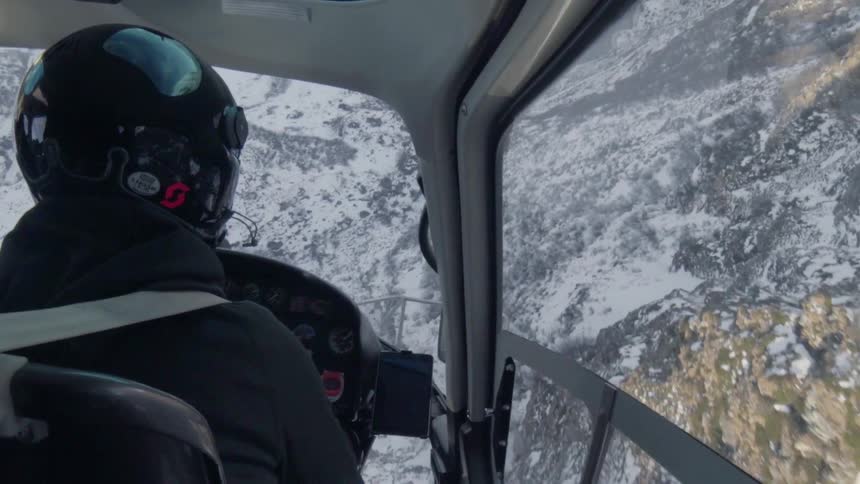 A pilot flies a helicopter over snowy mountains
