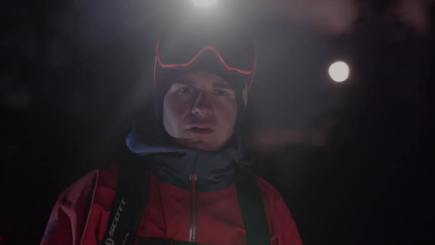 A man wearing snow gear and a headlamp removes his goggles