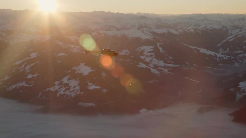 A helicopter flies over snow capped mountains on a sunny day
