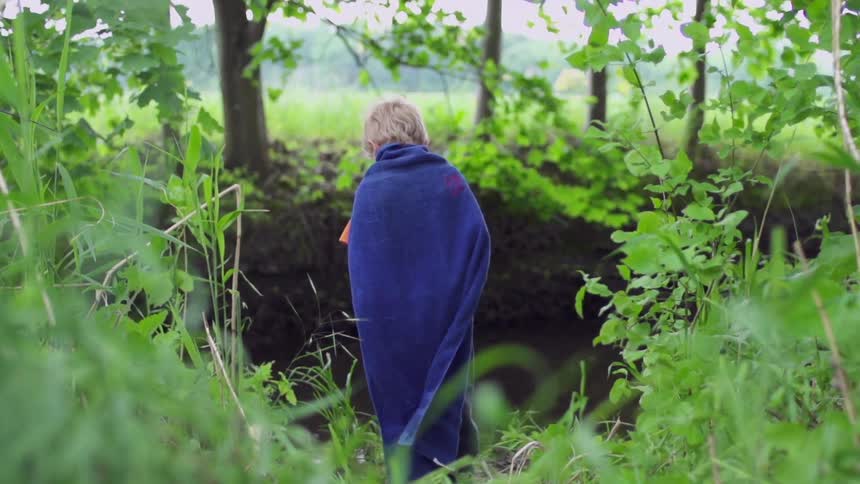 Child wearing a blue cape stands outside by trees