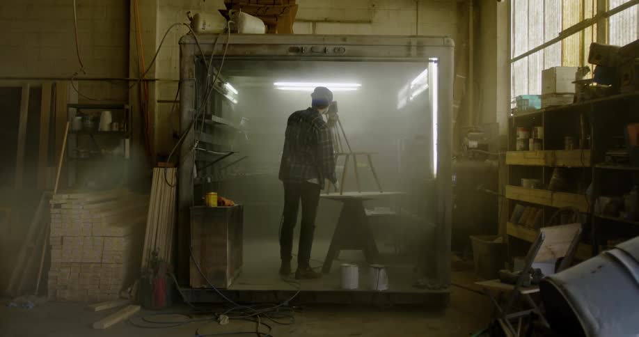 A man is spray painting a wooden chair in a room in a warehouse