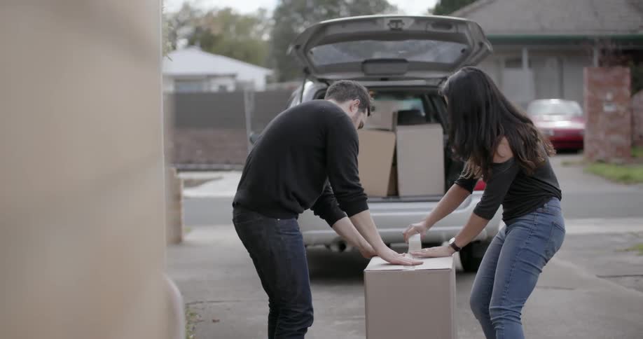 A man holds a box closed as a woman tapes it up in a driveway
