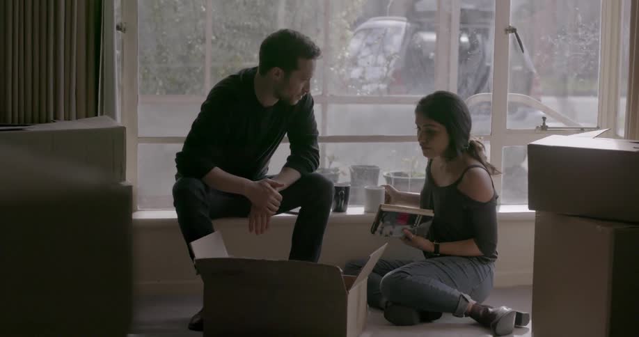 A man and woman look through a box of books as they sit in a room during the day