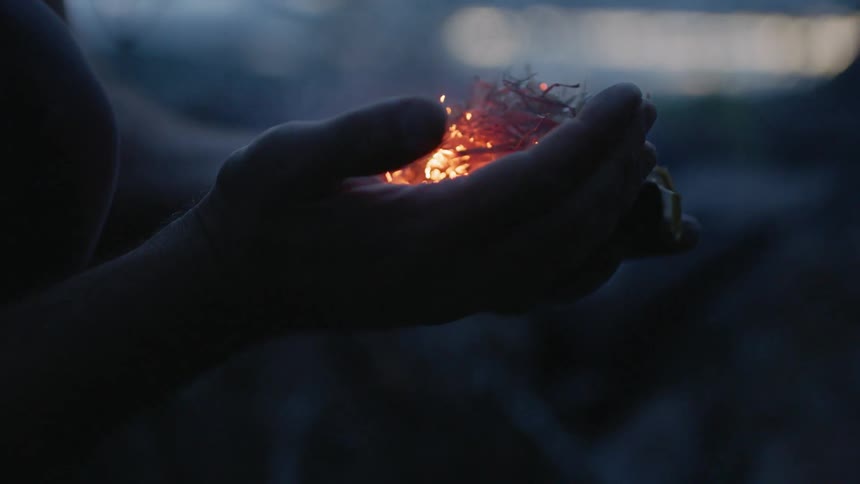 A person blows on some tinder in his hand to start a fire at dawn